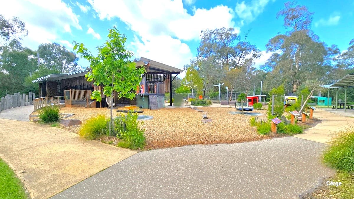 A building with a corrugated roof, slightly elevated. On the left, there's a ramp entry bordered by large stone structures and a metal railing. To the right, a concrete pathway leads to a shaded area with signages along the path. Trees and ornamental plants line the pathway's edge, with a few people moving about in the area.
