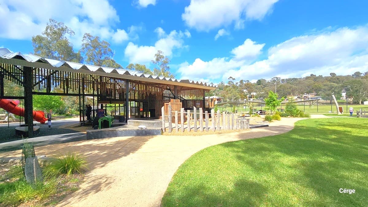 A building with a corrugated roof and wooden wall decorations. On the left, a small plant is barricaded with a chain fence. A stone structure fence is in the middle. To the left, there's a section of a long slide. On the right, a spacious grassy area extends. Further ahead, shaded areas with bench chairs for seating are visible. A few people are moving about.