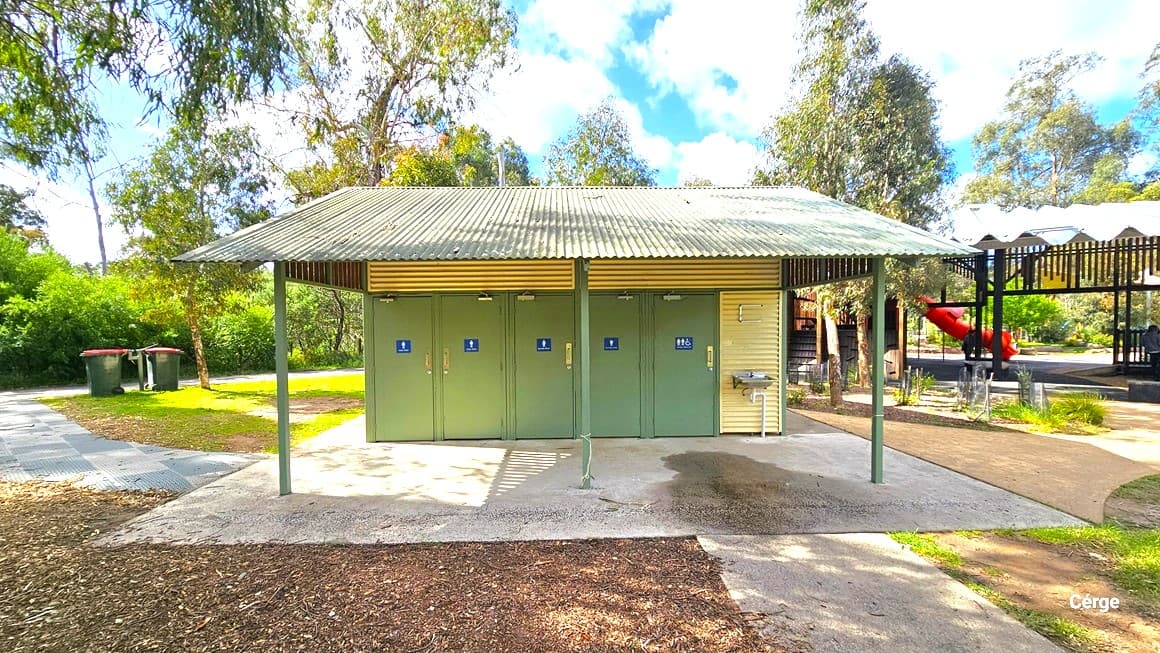 A toilet block with corrugated walls and roofing. There are 5 olive doors marked with gender specifications. A wash basin is on the right, and two green rubbish bins are on the left. To the right, a section of the building with a corrugated roof and a slide is visible. The ground is made of concrete, and colored tiles, and some areas are uneven with grass cover.