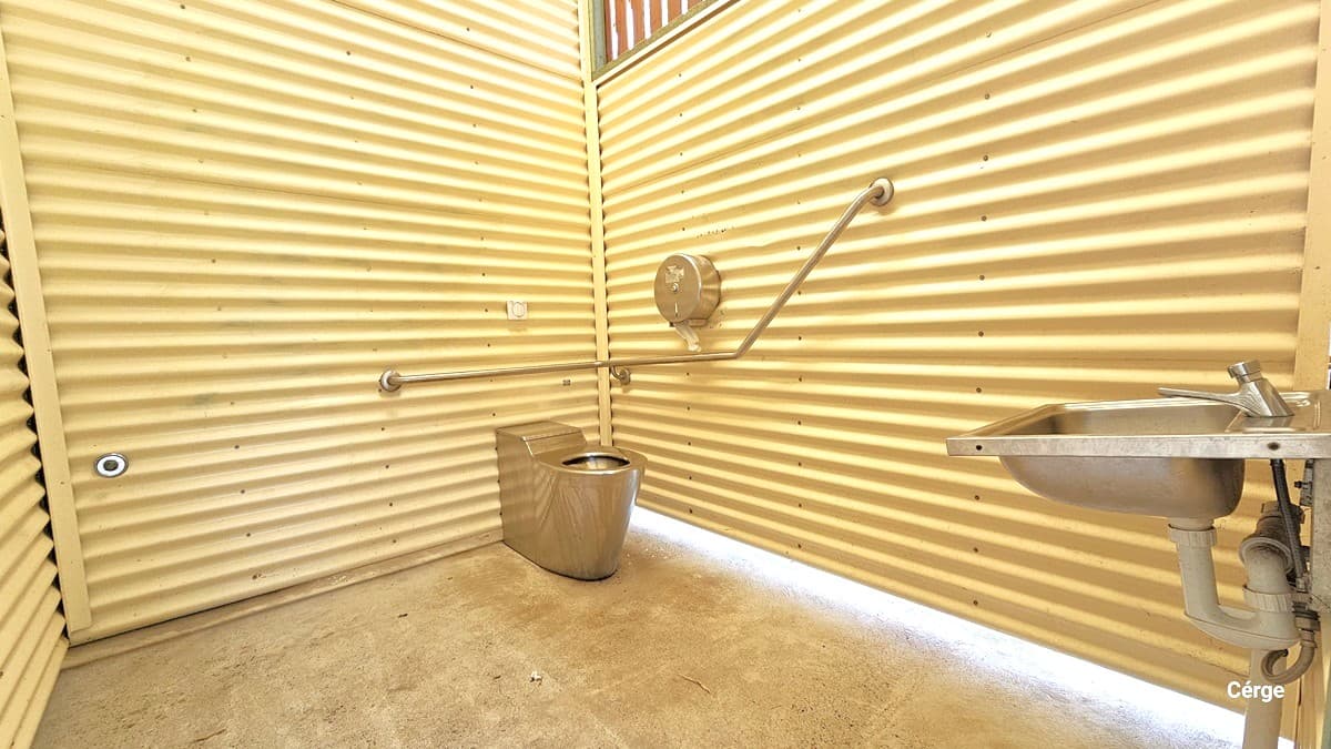 Within the bathroom, the interior wall features a cream-painted corrugated design. In one corner, there is a toilet bowl equipped with a handrail and a tissue dispenser. To the right, near the door, a washbasin is situated. The floor is made of concrete.