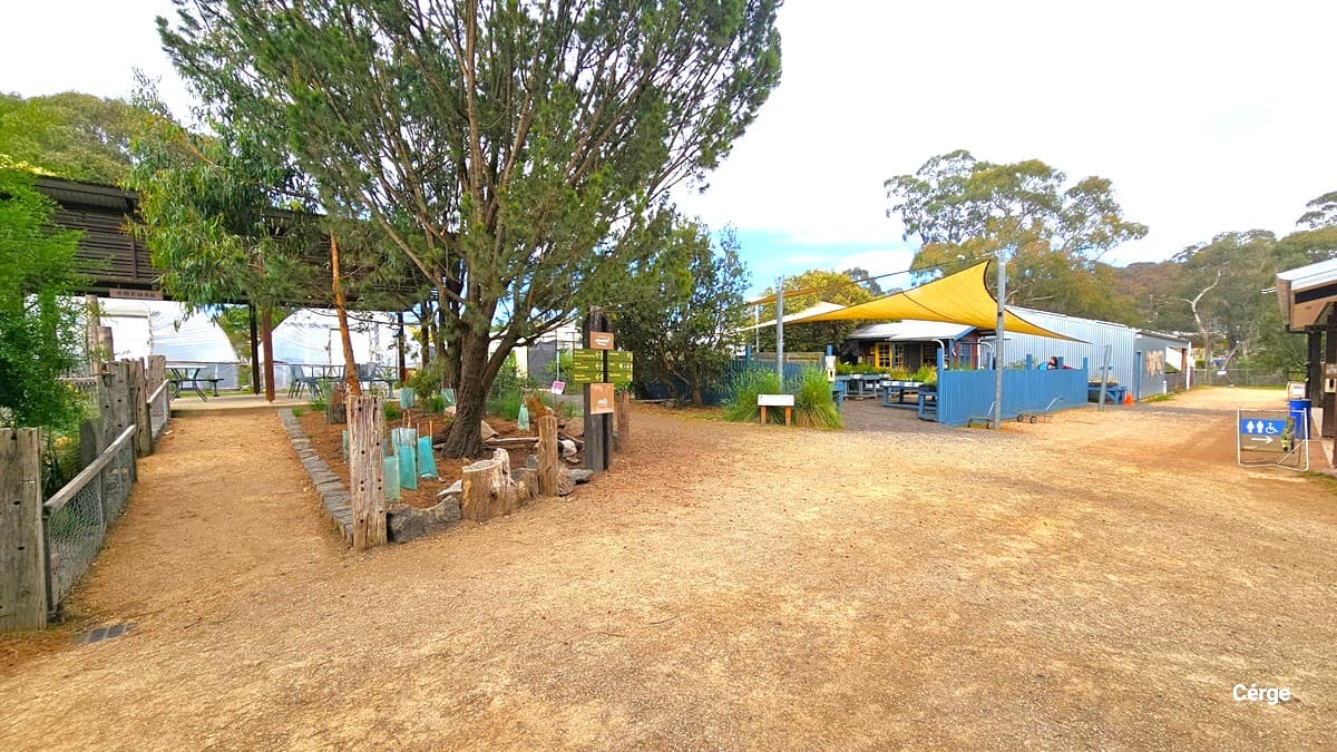 Another section of Edendale Community Farm is the nursery. This area comprises a spacious gravel expanse. On the left, a garden is delineated by stones and woods. A dirt pathway leads to a shaded seating area. In the center is the nursery, enclosed by a blue fence. Overhead, a yellow shade cloth provides shade for the plants underneath. To the right, a signage indicating accessible bathrooms is also present.