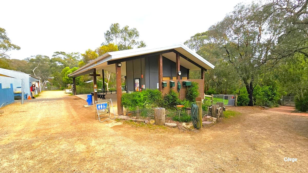 Adjacent to the nursery are the accessible bathrooms. A blue sign indicating the accessible bathrooms is displayed. On the right, a small garden landscape is situated, featuring bench chairs for seating. To the left, a gravel road leads to the opposite side of the garden, while on the right, another dirt road leads to gated areas.