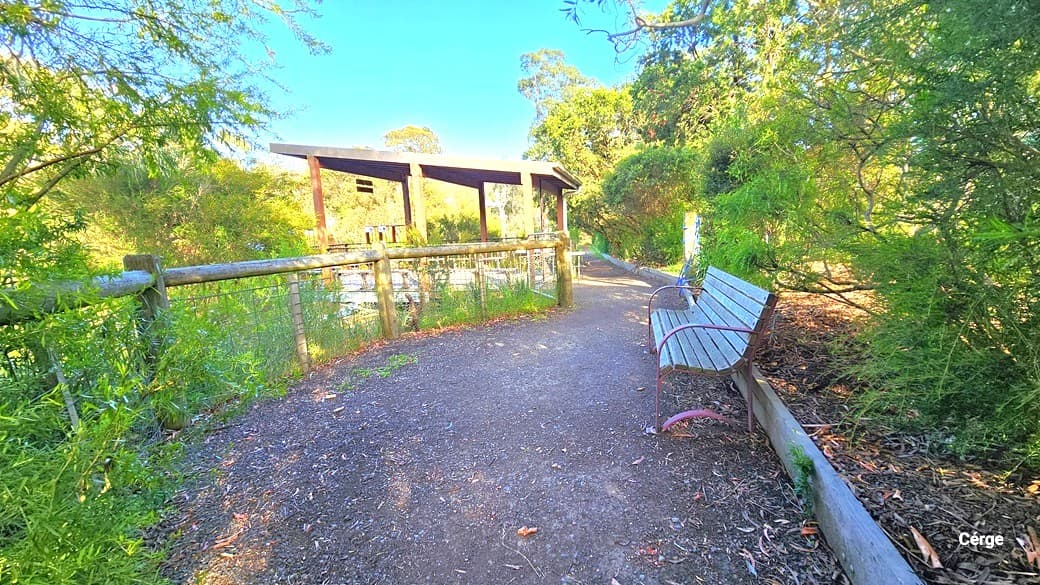 Proceeding along the gravel path, there's a section leading to the lake. A seating bench is on the right near the shrubs, while a low concrete and metal fence borders the left side. Further along, is the Dam Shelter area that offers a view of the lake. This area is surrounded by trees and shrubs.