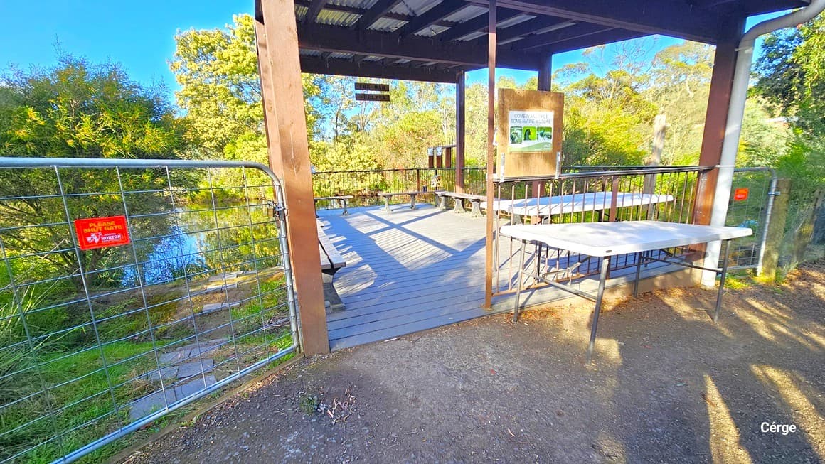 Continuing along the gravel path, is the Dam Shelter. This is a shaded area with a corrugated roof providing a view of the lake. Enclosed by an iron fence and gate, the space features bench chairs, white tables, and signage. A metal gate on the left reminds visitors to keep it closed. The area is surrounded by grass, shrubs, and tall mature trees.