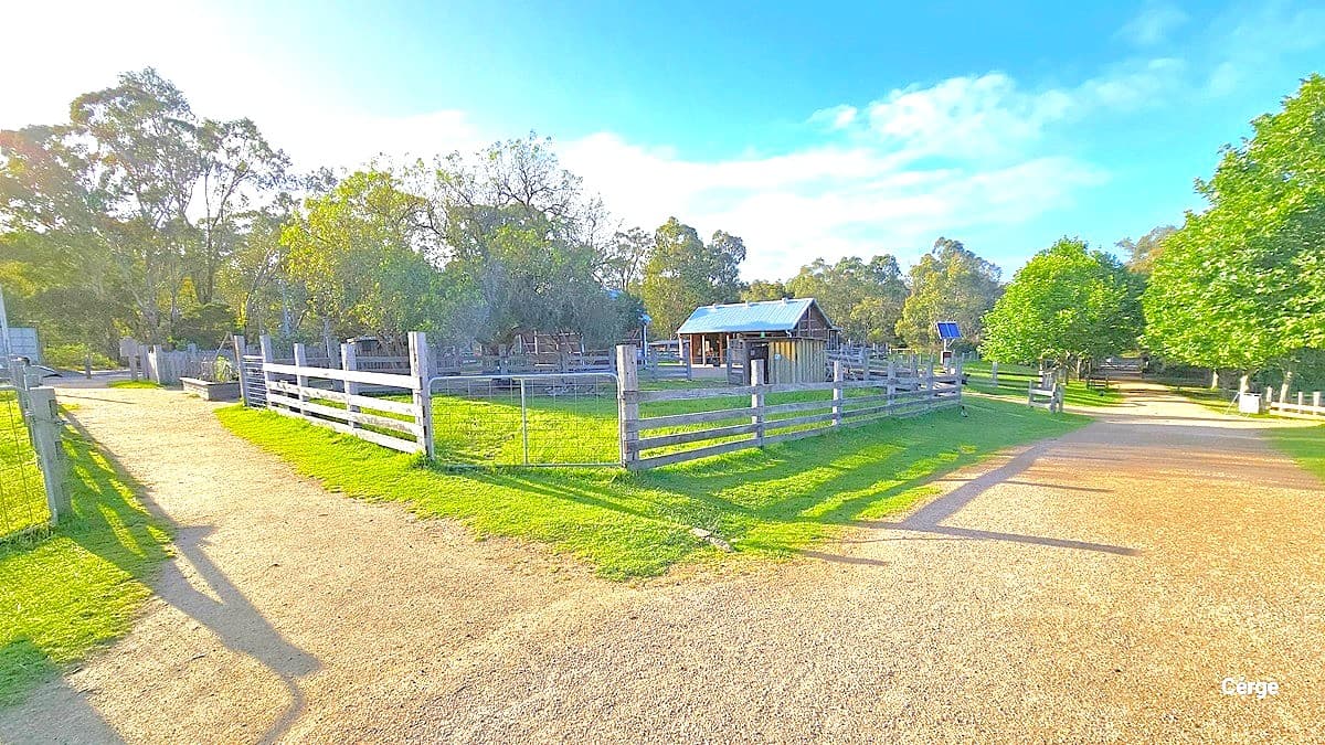 Two gravel pathways leading in different directions. To the left is a grassy area enclosed with a concrete and metal fence. In the middle is another enclosed grassy area with farm sheds. Continuing along the pathway on the right, there is a solar street light. Trees and a wooden fence border the pathway.