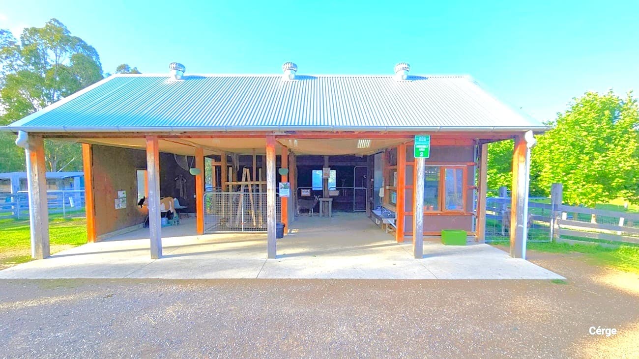 This part of Edendale Farm features a farm shed with a corrugated design roof. Three wind-driven stainless steel fans are installed on the roof. Farm equipment, including a goal model, is in place. Multiple banners are affixed to the wall. A wash area is also located on the right side near the glass window. The area is surrounded by grass and a fence.