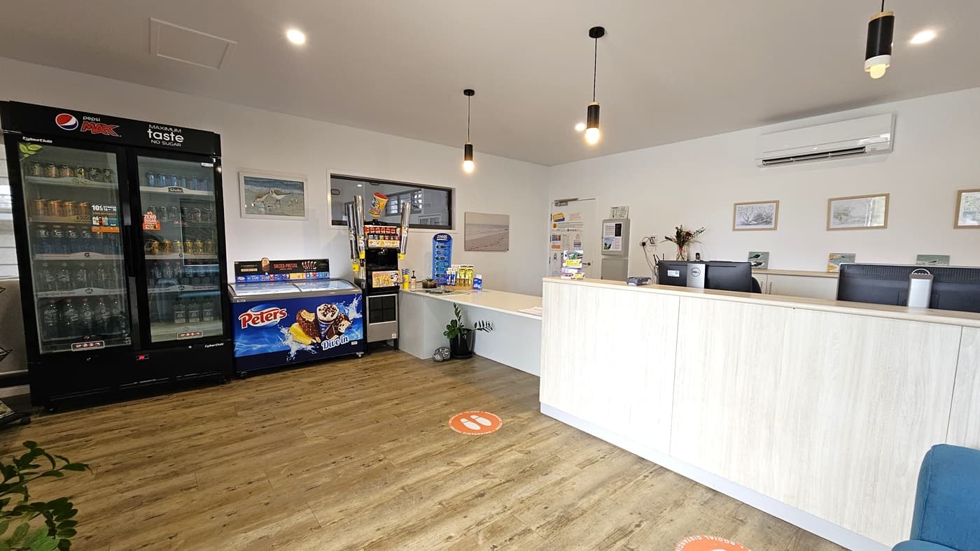 A reception desk with a timber front holds computers on the right. Adjacent is a lower desk with brochures. To the left is a fridge displaying cold drinks and an ice cream freezer, along with a drinks dispenser. The white ceiling and wall feature light fixtures, posters, and decorations. The timber-tiled floor is decorated with a potted plant on the left, and a blue couch is positioned in front of the reception desk.