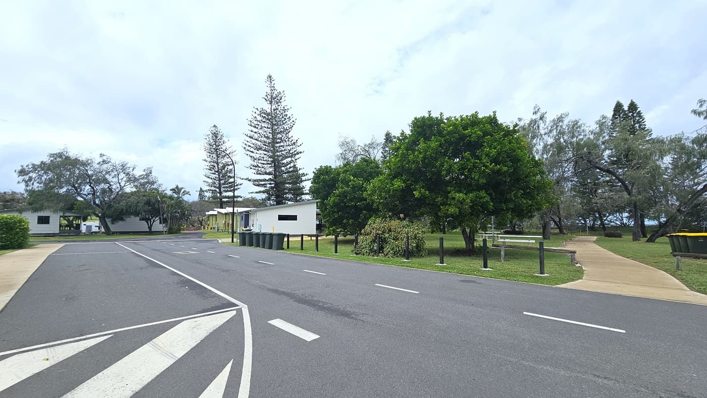 The road features white road markings, bordered by black bollards. A concrete pathway leads to the park on the right, amidst trees and grass. Rubbish bins line the road's edge. In the distance, cabins and a tall street light are visible.