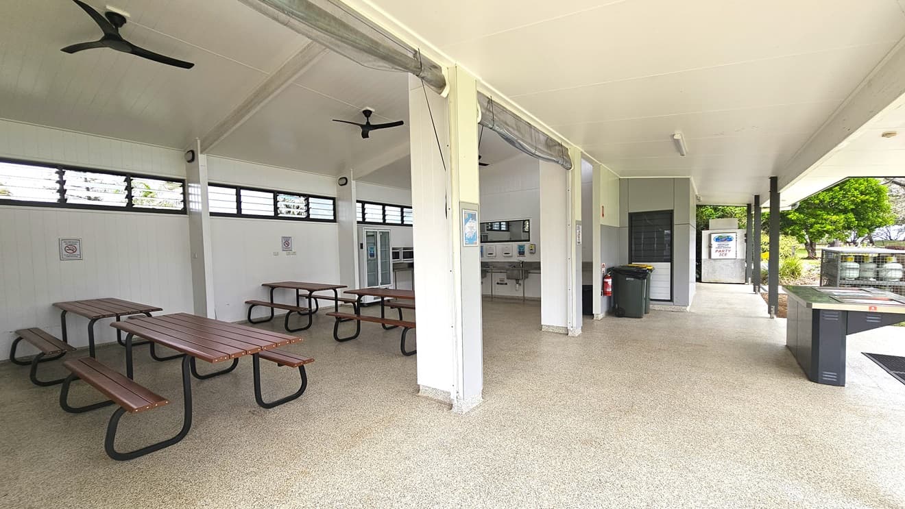The BBQ area is spacious with picnic tables on the sides and a fridge. A sink and mirror are in the middle wall, and to the right is a barbecue grill. Gas tanks are in a cage nearby, with two rubbish bins and a fire extinguisher on the outer wall. White ceilings and walls feature ceiling fans and jalousy windows for ventilation. Safety reminders are affixed to the walls. Trees are visible on the right.