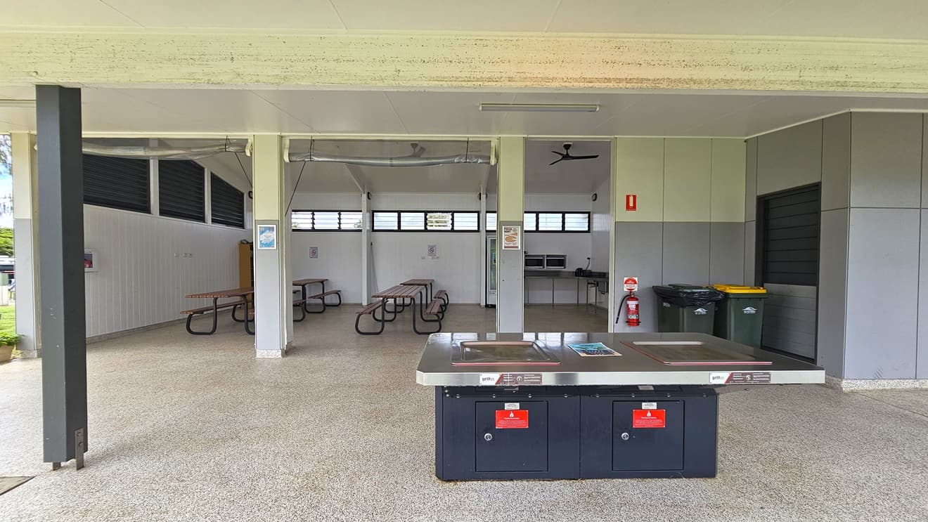 The spacious BBQ area has picnic tables and a wooden display rack in the left corner. On the right, there's a fridge, microwave ovens, and a sink. The pitched white ceiling hosts ceiling fans. Walls showcase a white timber design, with jalousy windows and light fixtures. Safety reminders are affixed to walls. In front is a barbecue grill with safety warning stickers. Two rubbish bins and a fire extinguisher are positioned on the outer wall on the right.