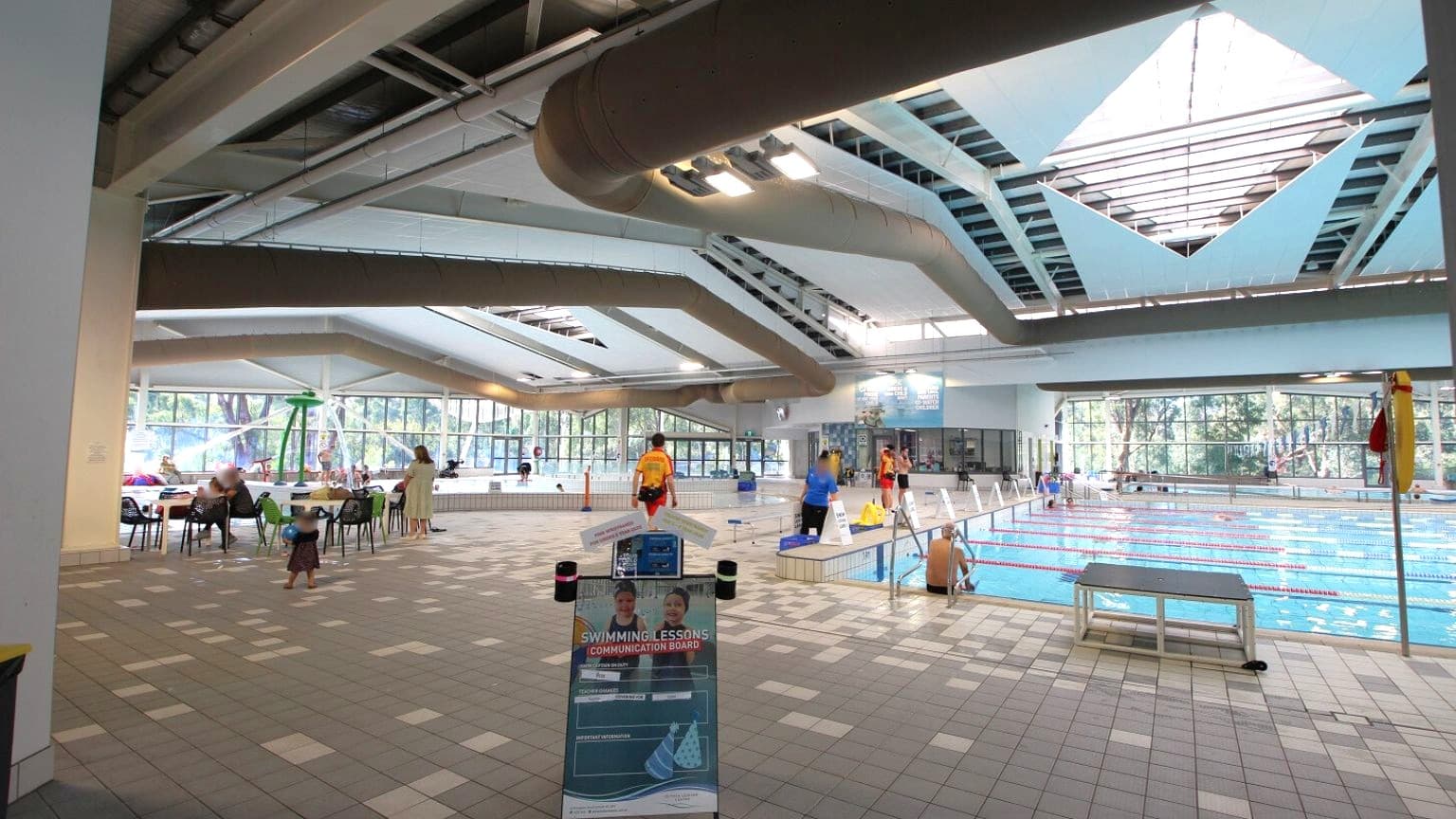 An indoor swimming pool area with a tiled floor. On the left, there are tables and chairs with people sitting and a child walking. In the centre, a sign about swimming lessons is visible. On the right, the pool has lane dividers and a few people standing by the edge. The ceiling features large, geometric skylights, and the background shows large windows with a view of trees outside.