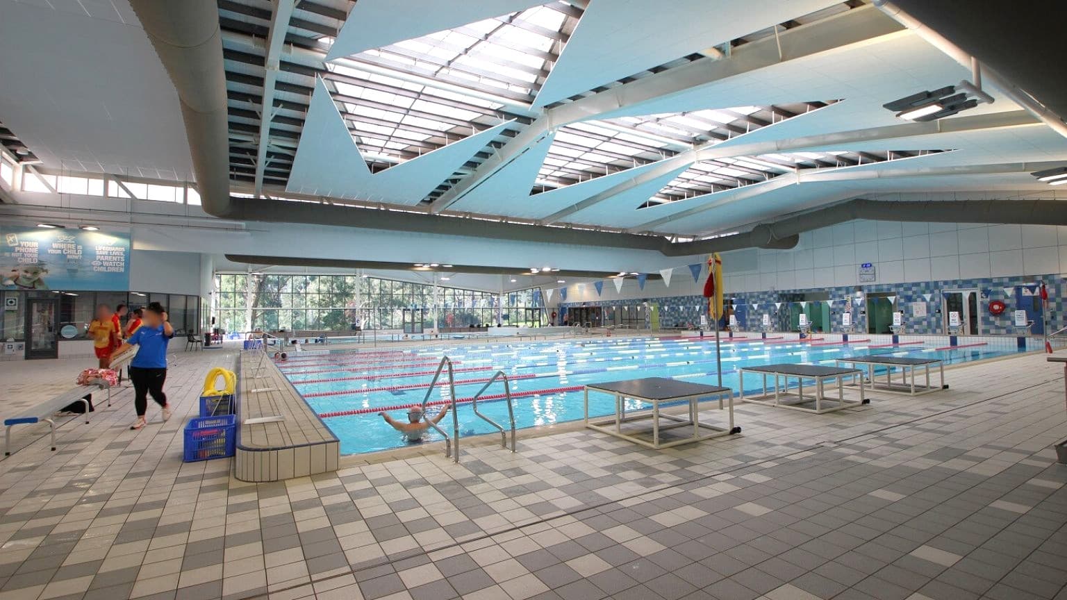 An indoor swimming pool with multiple lanes, surrounded by a tiled deck. On the left, a few people are walking near the poolside, with some equipment like a yellow lifebuoy and a blue crate nearby. The ceiling features large, angular skylights allowing natural light to illuminate the space. The walls have large windows and some signage. The floor is covered with multicoloured tiles, and there are diving platforms on the right side of the pool.