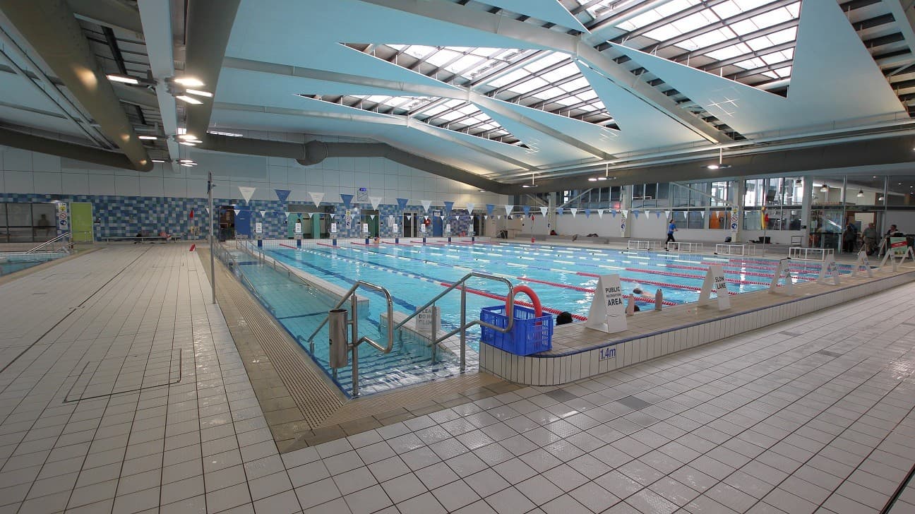 Indoor swimming pool with multiple lanes, surrounded by a tiled deck. On the left, there's a wide walkway with a non-slip surface. The right side features a row of windows and seating areas. The ceiling has large skylights allowing natural light to illuminate the space. Safety signs and pool equipment are visible around the pool's edge.