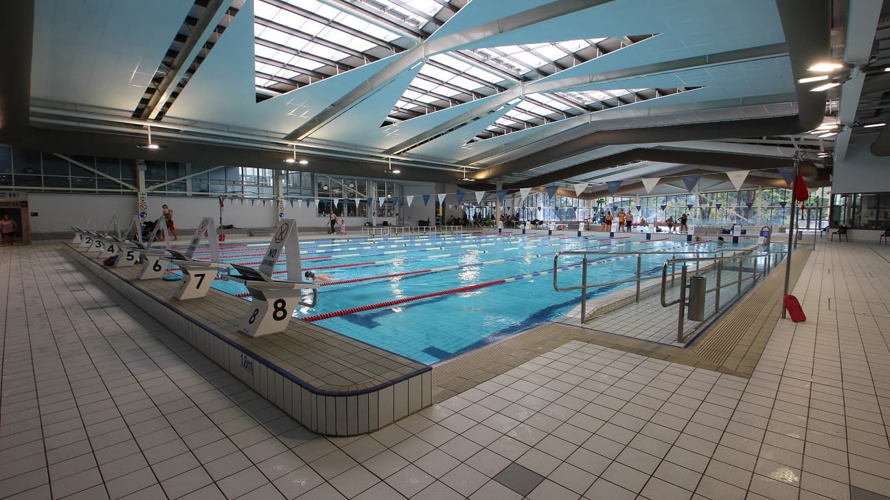Indoor swimming pool with multiple lanes, each marked with numbers on starting blocks. The pool is surrounded by a tiled deck. The ceiling features large skylights and angular beams, allowing natural light to illuminate the area. On the right, there are handrails leading into the pool, and a few people are visible swimming and standing by the poolside. The background shows large windows and some seating areas.
