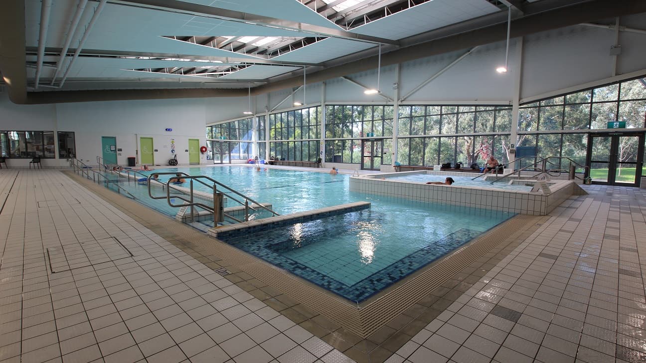 Indoor swimming pool with a tiled deck surrounding it. The pool has metal handrails and a few people are in the water. Large windows on the right side provide a view of trees outside. The ceiling is high with exposed beams and skylights. The floor is covered with light-coloured tiles. Doors and emergency equipment are visible on the far wall.