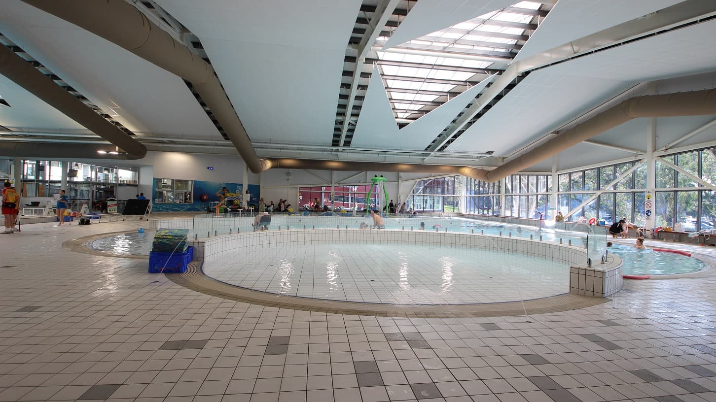 A spacious indoor swimming pool area with a large, circular shallow pool in the centre. The floor is tiled with a mix of beige and grey tiles. On the left, there are people standing near the pool's edge. The right side features large windows allowing natural light to enter, with trees visible outside. The ceiling is high with geometric designs and skylights. In the background, there are more pools and people, along with some poolside equipment.