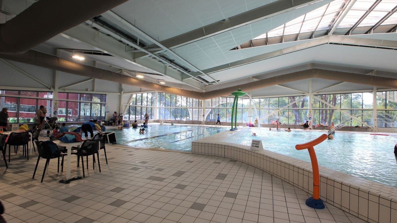 Indoor swimming pool area with a tiled floor. On the left, there are chairs and tables with people sitting and standing nearby. The pool is in the centre, with several people swimming and playing. A green water feature stands in the pool. Large windows in the background allow natural light to enter, and trees are visible outside. The ceiling is high with exposed beams and lighting fixtures.