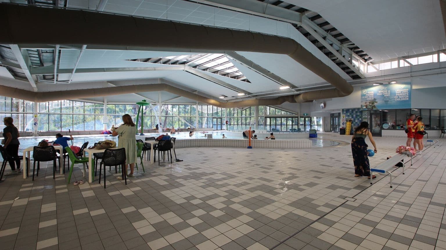 Indoor swimming pool area with a tiled floor in shades of green and beige. On the left, several people are seated at tables with chairs, some watching the pool. In the centre, a pool with swimmers and a lifeguard is visible. On the right, a person is bending over near a small poolside structure, and a group of people are gathered near the wall. The ceiling has a geometric design with skylights, and large windows in the background allow natural light in.