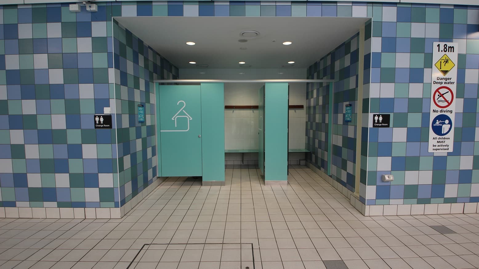 A changing room entrance with three turquoise doors, one slightly open, revealing benches and hooks inside. The walls are covered in a blue and white checkered tile pattern. On the right, there are signs indicating a depth warning, no diving, and supervision requirements. The floor is tiled in a light beige colour, and the ceiling has recessed lighting.