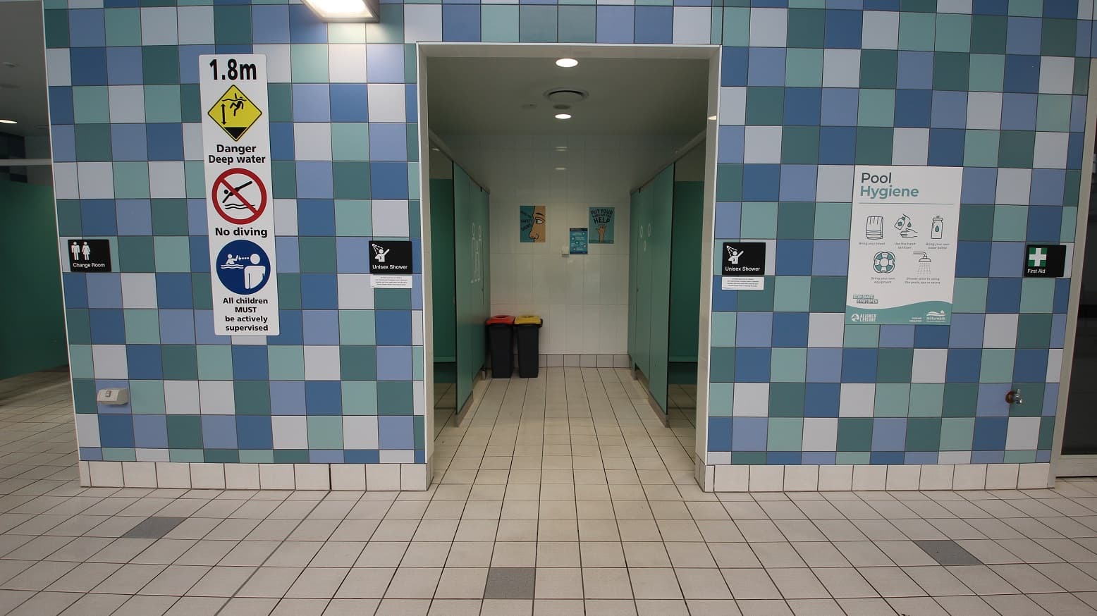 A tiled wall with a blue and white checkerboard pattern. In the centre, there's an entrance to a shower area with two bins inside. Above the entrance, signs indicate "1.8m Danger Deep Water," "No Diving," and "All children MUST be actively supervised." On the left, a sign for the "Change Room" and on the right, a "Pool Hygiene" poster and a "First Aid" sign. The floor is tiled in a light colour, matching the wall's pattern.