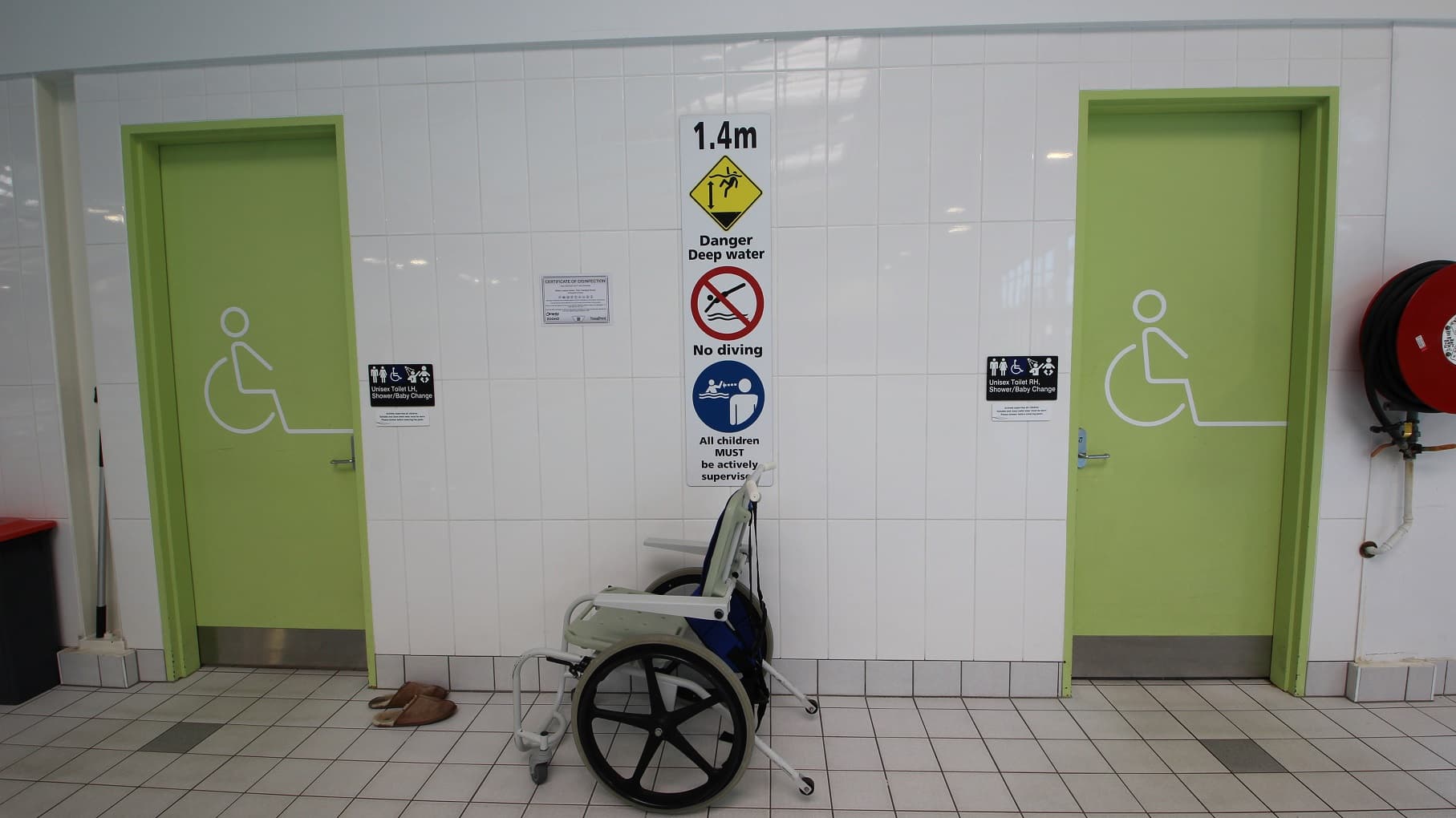 Two light green doors with wheelchair symbols are on either side of a white-tiled wall. Between the doors, a sign indicates a water depth of 1.4 metres, warning of deep water and prohibiting diving. Below the sign, a wheelchair is positioned against the wall. A pair of brown sandals is on the floor near the left door. A red fire hose reel is mounted on the wall to the right. The floor is tiled in a light grey pattern.