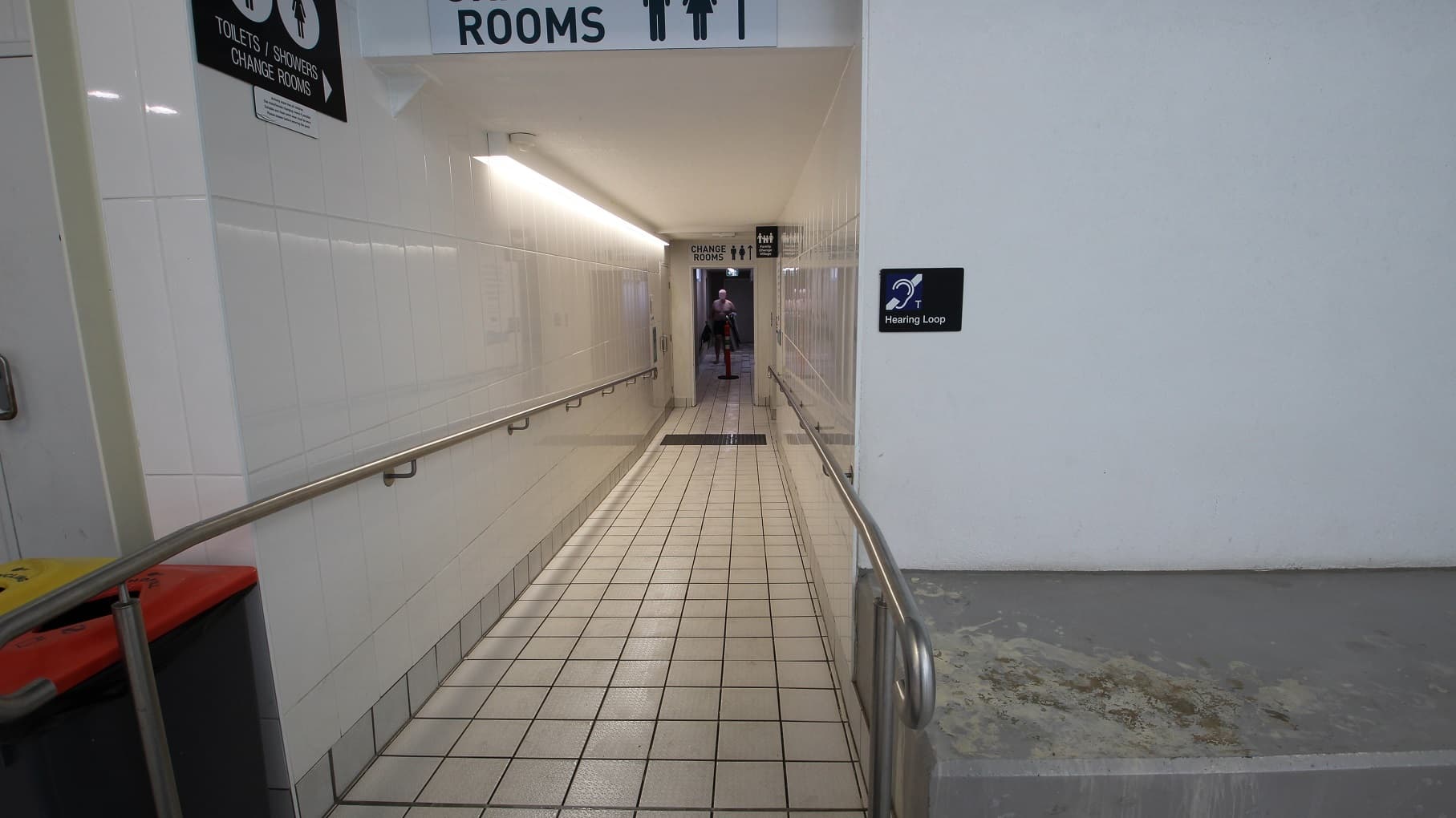 A narrow corridor with tiled walls and floor, leading to changing rooms. On the left, there's a handrail and a bin with a red lid. On the right, a sign indicates a hearing loop. Overhead, signs direct to toilets, showers, and changing rooms. At the end of the corridor, a person is visible in the distance. The ceiling is plain with fluorescent lighting.