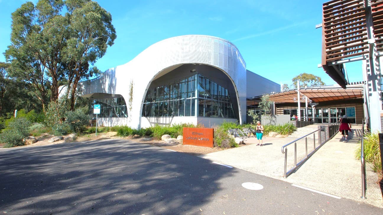 Modern building with a curved, perforated metal facade on the left and a structure with horizontal wooden slats on the right. In the centre, a pathway leads to the entrance, flanked by greenery and a sign reading "Eltham Leisure Centre". The ground is paved, and there is a ramp on the right side. The building features large glass windows, and there are trees visible in the background.