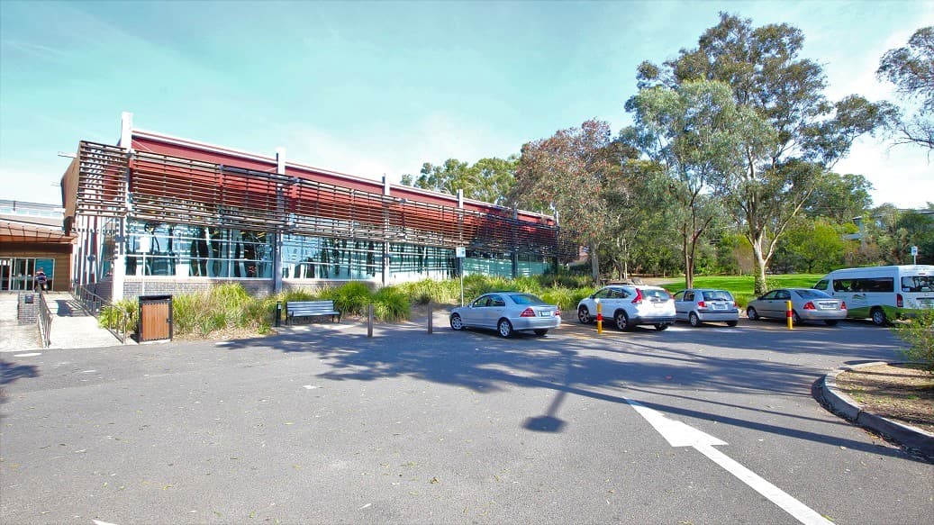 A modern building with a curved, metallic facade and large glass windows is on the left. In front, there's a sign with text and landscaped greenery. A pathway leads to the entrance on the right, where people are walking. The building is surrounded by tall trees, and there's a clear blue sky above. A railing lines the path, and the ground is a mix of pavement and gravel.