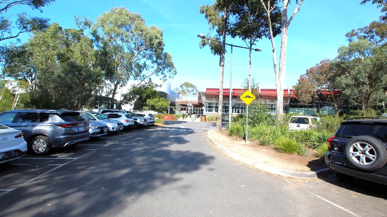 A modern building with a facade featuring horizontal slats and large glass windows is on the left. In front, there is a small parking area with several cars and a minibus. The ground is paved with asphalt, and there are some bollards near the parking spaces. On the right, there are tall trees and green grass, suggesting a park or garden area. The sky is clear, and the scene is well-lit, indicating daytime.