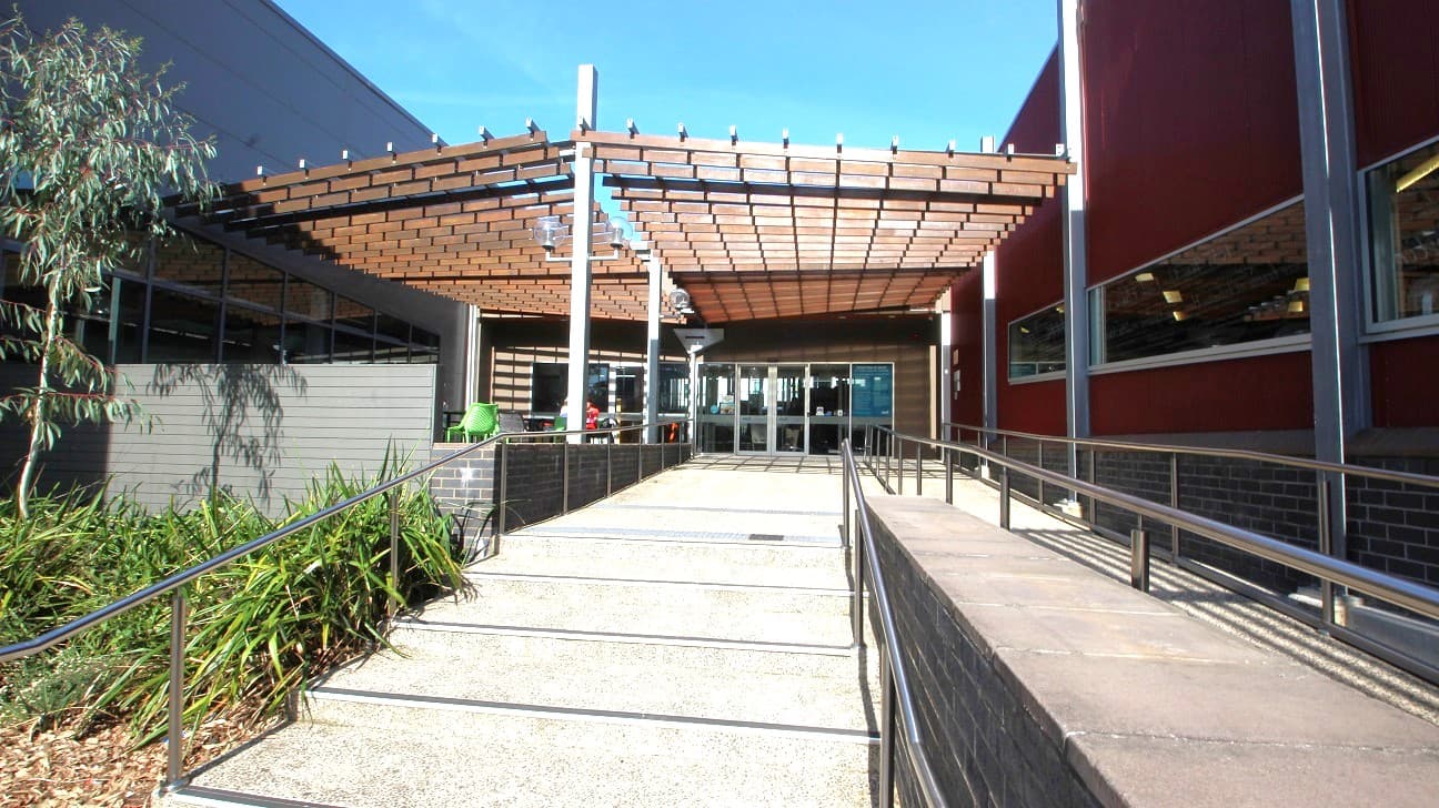 A car park with several vehicles lined up on the left side, including a grey SUV and white cars. On the right, a yellow road sign is visible near some greenery. In the background, a building with a red roof and large windows is partially obscured by trees. The road curves slightly, and tall trees are scattered throughout the area. The sky is clear and blue.