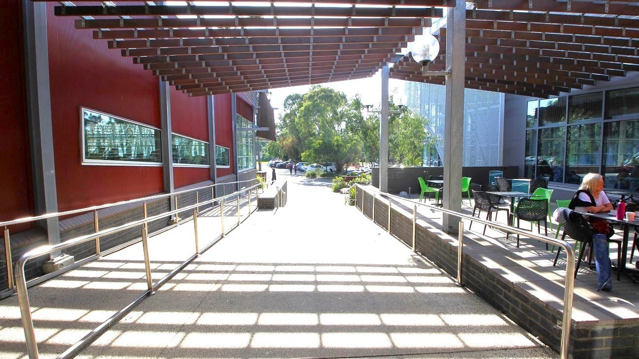 A pathway with metal railings runs through the centre, leading to an outdoor area with trees and parked cars in the background. On the left, a red building with long horizontal windows. On the right, a seating area with tables and chairs, where a person is sitting. Above, a grid-like structure provides partial shade, casting shadows on the ground.