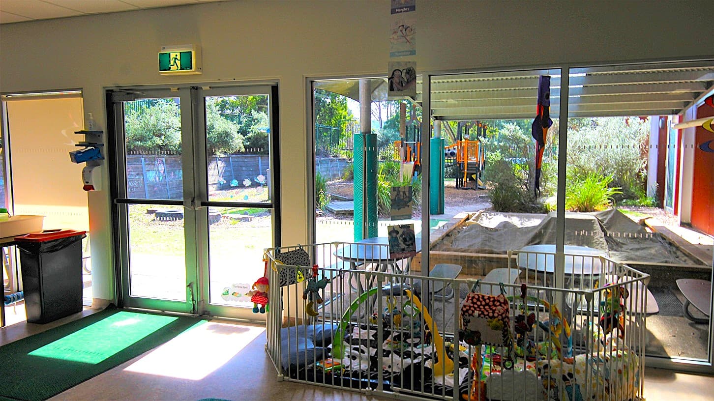 White fence at the entrance Child Care Centre at Eltham Leisure Centre. Beyond the fence are toys, costumes, kids' decorations, kids' drawings and paintings, and a soft green mat to protect children during playing.