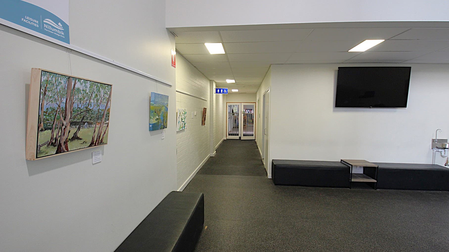 A small space with TV, paintings, and black sofa chairs in the white corridor at the entrance of Child Care at Eltham Leisure Centre. Underneath the TV is a faucet and between the sofa chairs is a magazine table.