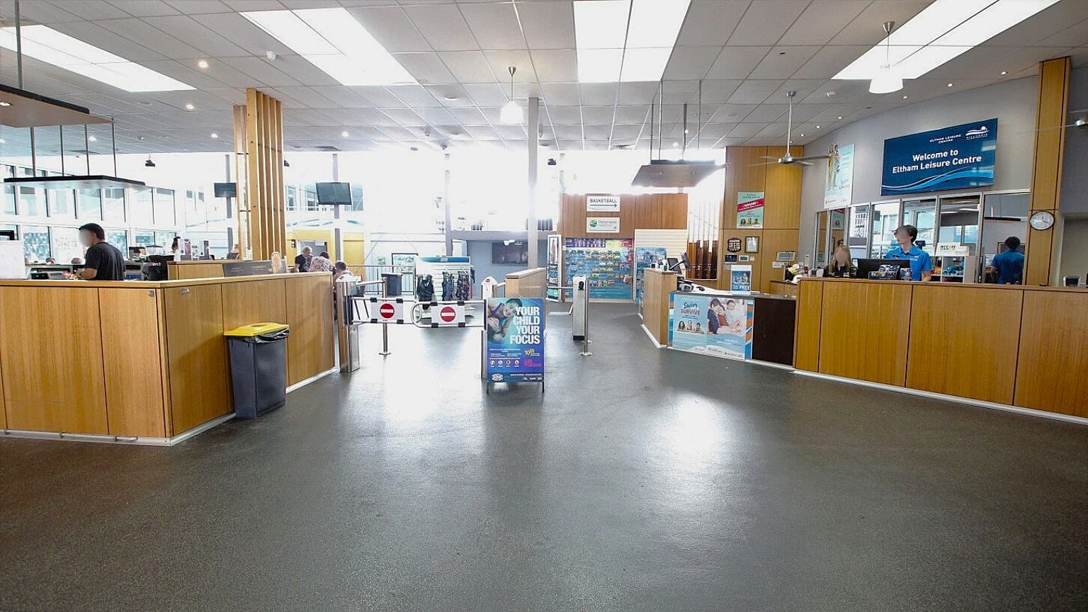 A spacious interior with a dark floor and a high ceiling featuring large rectangular lights. On the left, a wooden counter with a yellow-lidded bin and a person behind it. In the centre, a sign and a barrier with "no entry" symbols. On the right, another wooden counter with two people behind it, and a digital sign above reading "Welcome to Eltham Leisure Centre." The background has glass panels and more signage.