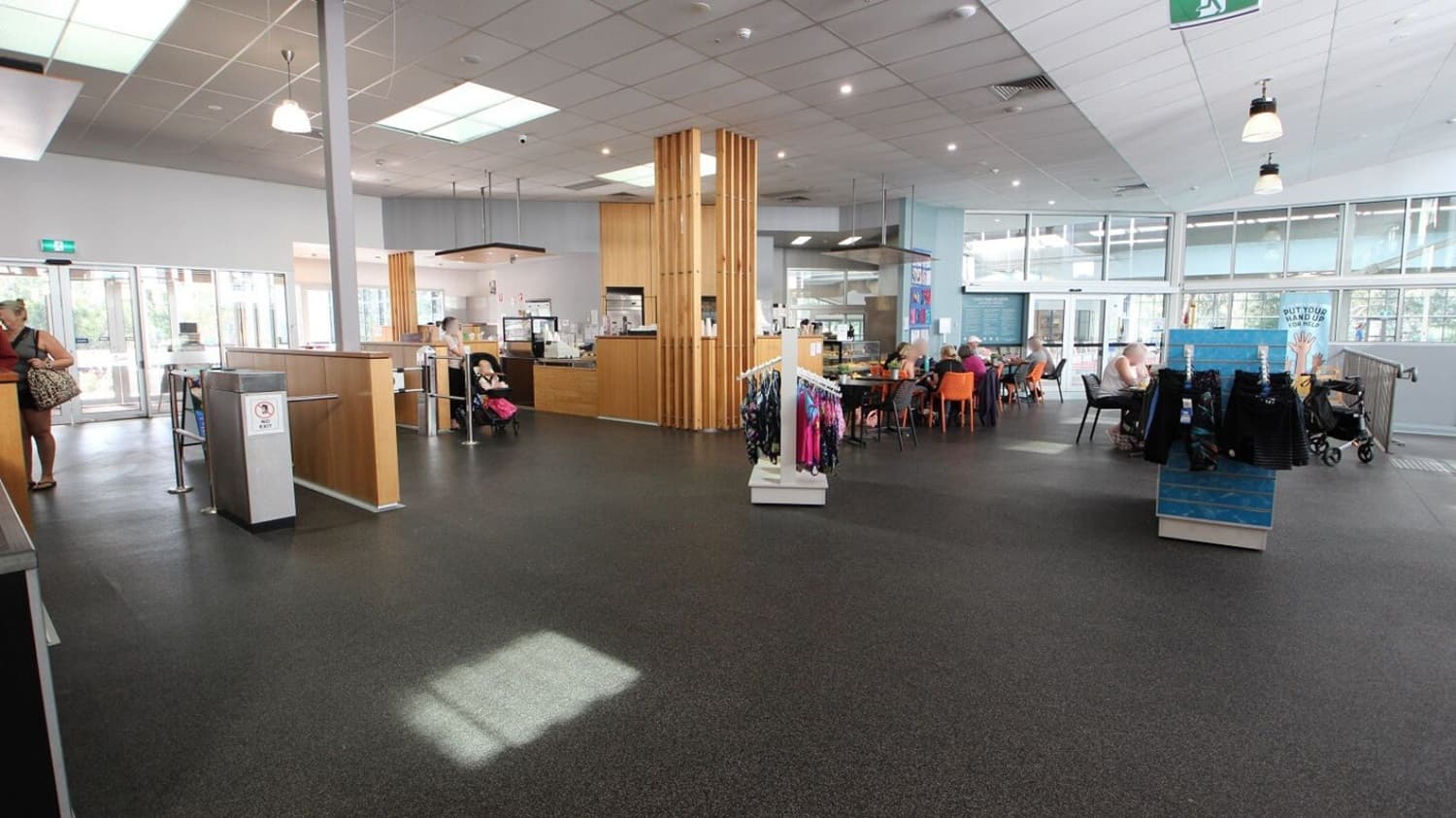 A spacious indoor area with a dark floor and a ceiling featuring recessed lighting and skylights. On the left, a woman stands near a counter with a glass entrance behind her. In the centre, a wooden column and a small café area with people seated. On the right, a display rack with clothes and a group of people sitting at tables with orange chairs. Large windows line the background, allowing natural light to fill the space.