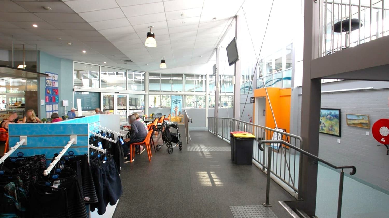 A spacious indoor area with a high ceiling and large windows on the right, allowing natural light to flood in. On the left, there are people seated at tables with orange chairs, and a blue partition with hanging items. In the middle, a pram is parked near the tables. The floor is dark and textured, with a ramp and railing leading to a higher level. On the right wall, there are paintings and a red fire hose reel. The ceiling has several hanging lights.
