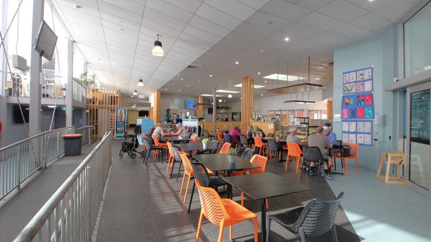 A spacious indoor café area with a high ceiling and modern design. On the left, a metal railing lines a walkway with a mounted TV screen above. The floor is a mix of dark and light tiles. In the centre, there are black tables with orange and black woven chairs. People are seated, engaged in conversation. The right side features a light blue wall adorned with colourful artwork. The café counter is visible in the background, with hanging lights and a menu display.