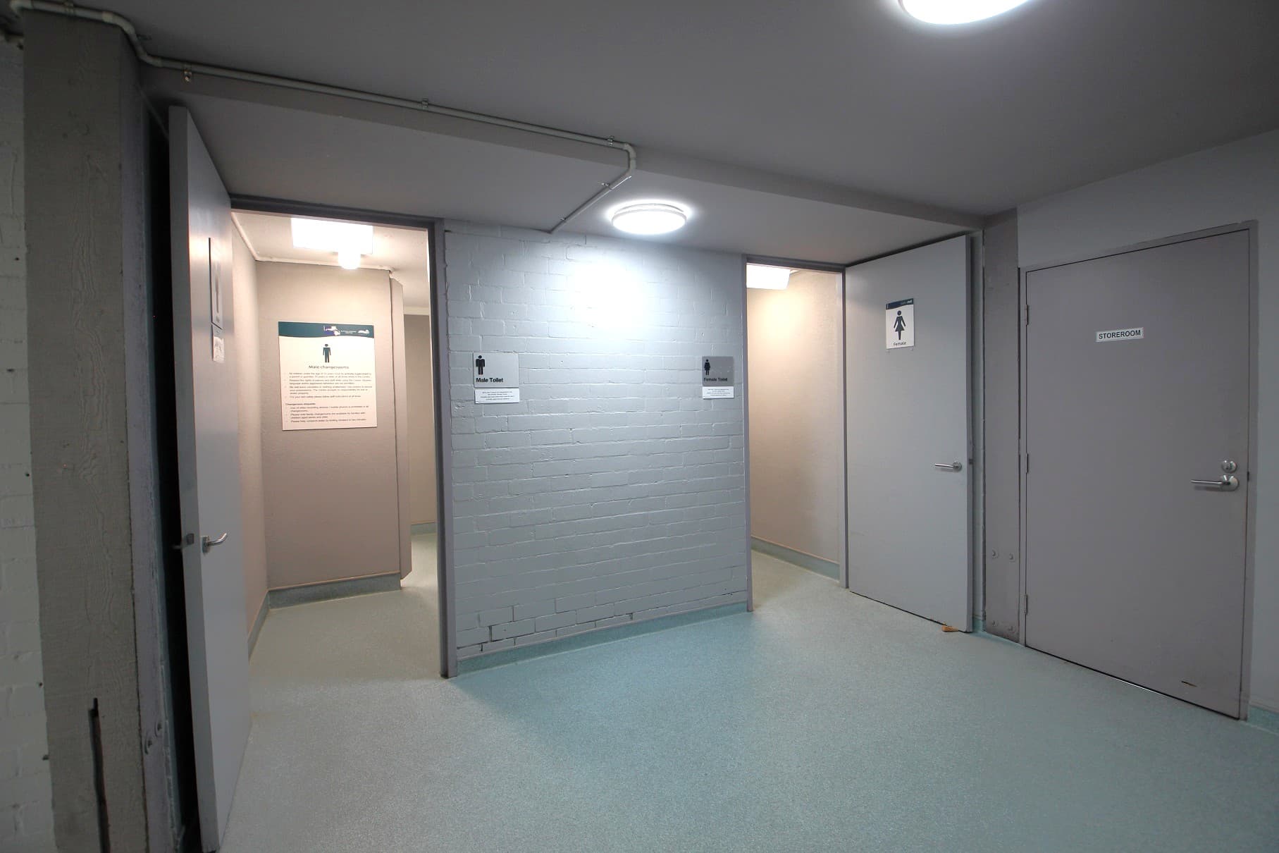 A corridor with three doors. On the left, a door is open, leading to a room with a sign indicating a male toilet. In the centre, a wall with a sign for male toilets. On the right, two closed doors; one marked with a female toilet sign and the other labelled "Storeroom". The floor is light-coloured, and the ceiling has round, recessed lights.
