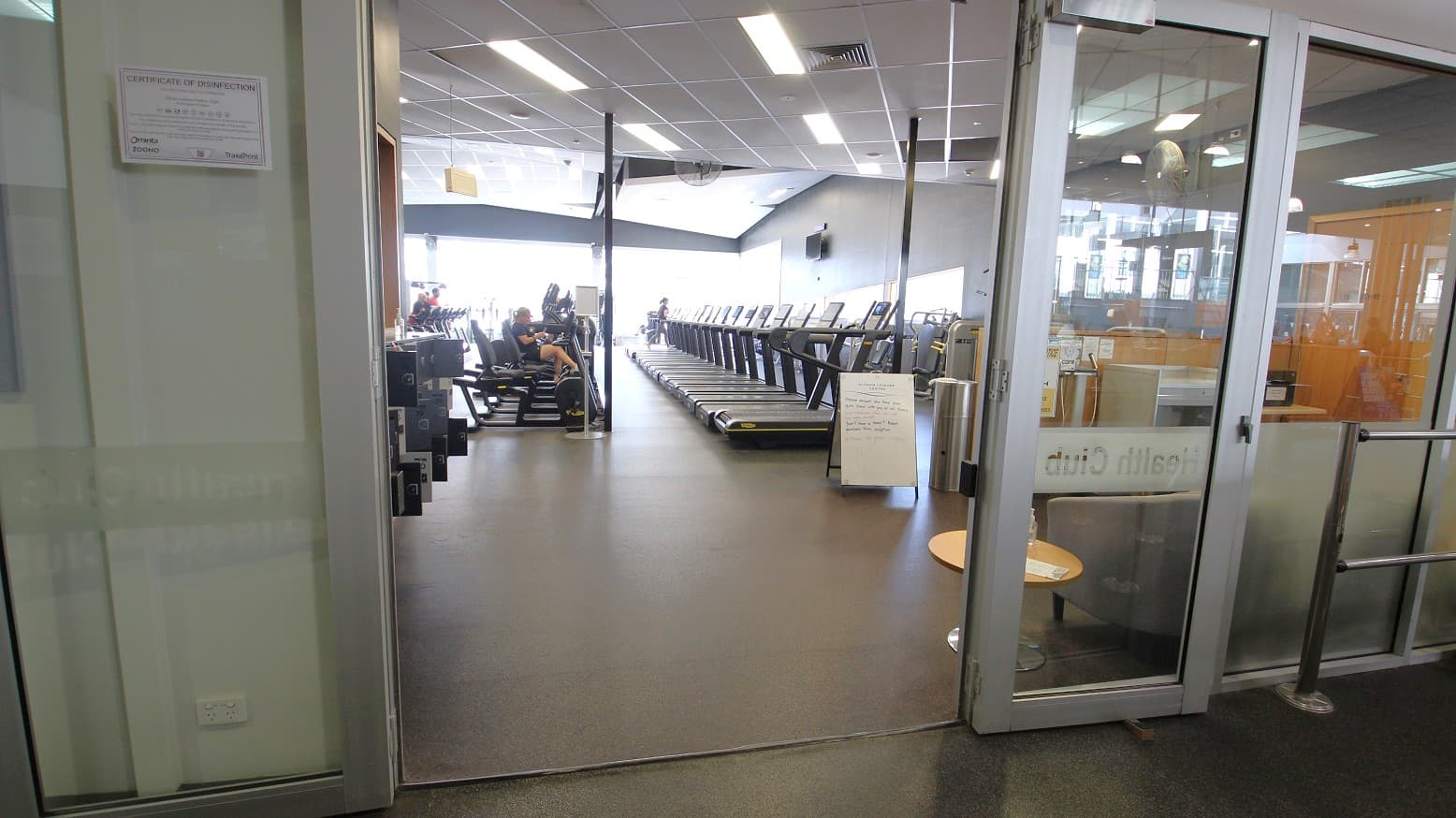 A disinfection certificate is mounted at the left side of the main entrance door of the Gymnasium at Eltham Leisure Centre. Several treadmills are lined directly at the main entrance.