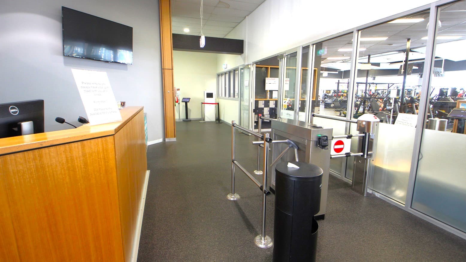 A timber desk and turnstile gates at the entrance of the Gymnasium at Eltham Leisure Centre. The gymnasium has glass walls that entice you with different pieces of equipment available for you to try.