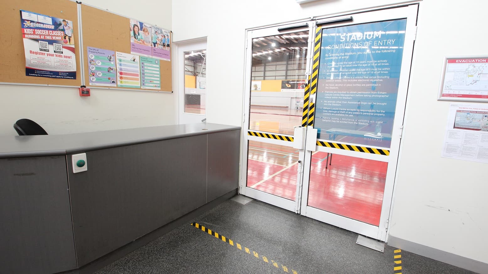 Adjacent to the dark grey reception desk at the entrance of Eltham Leisure Centre is the glass door with yellow and black safety tape. On the right side of the door is an evacuation map.