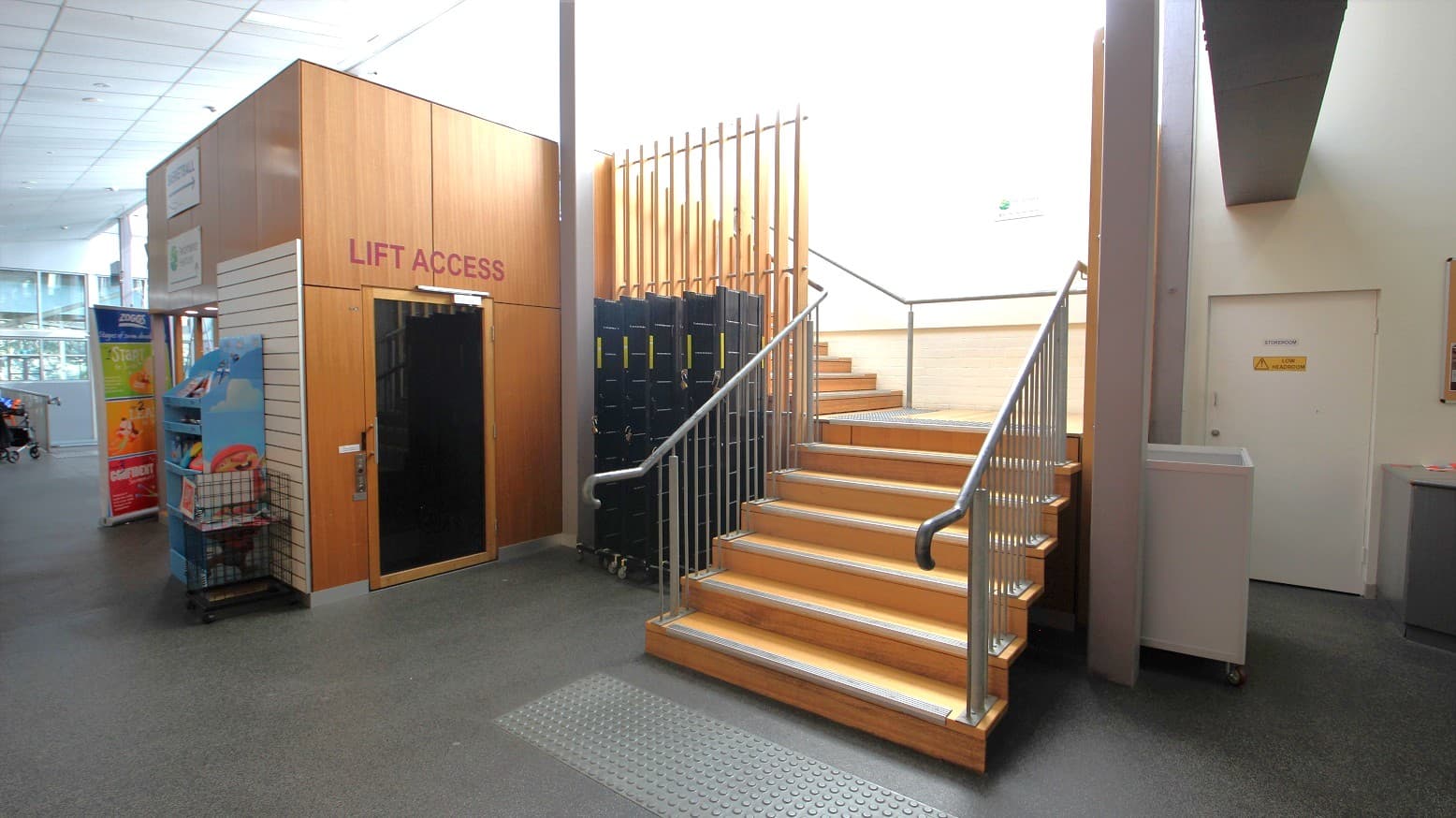 One elevator with a “lift access” sign and a single staircase at the Yoga studio in Eltham Leisure Centre. The staircase has grey metal railings and is beside the storeroom