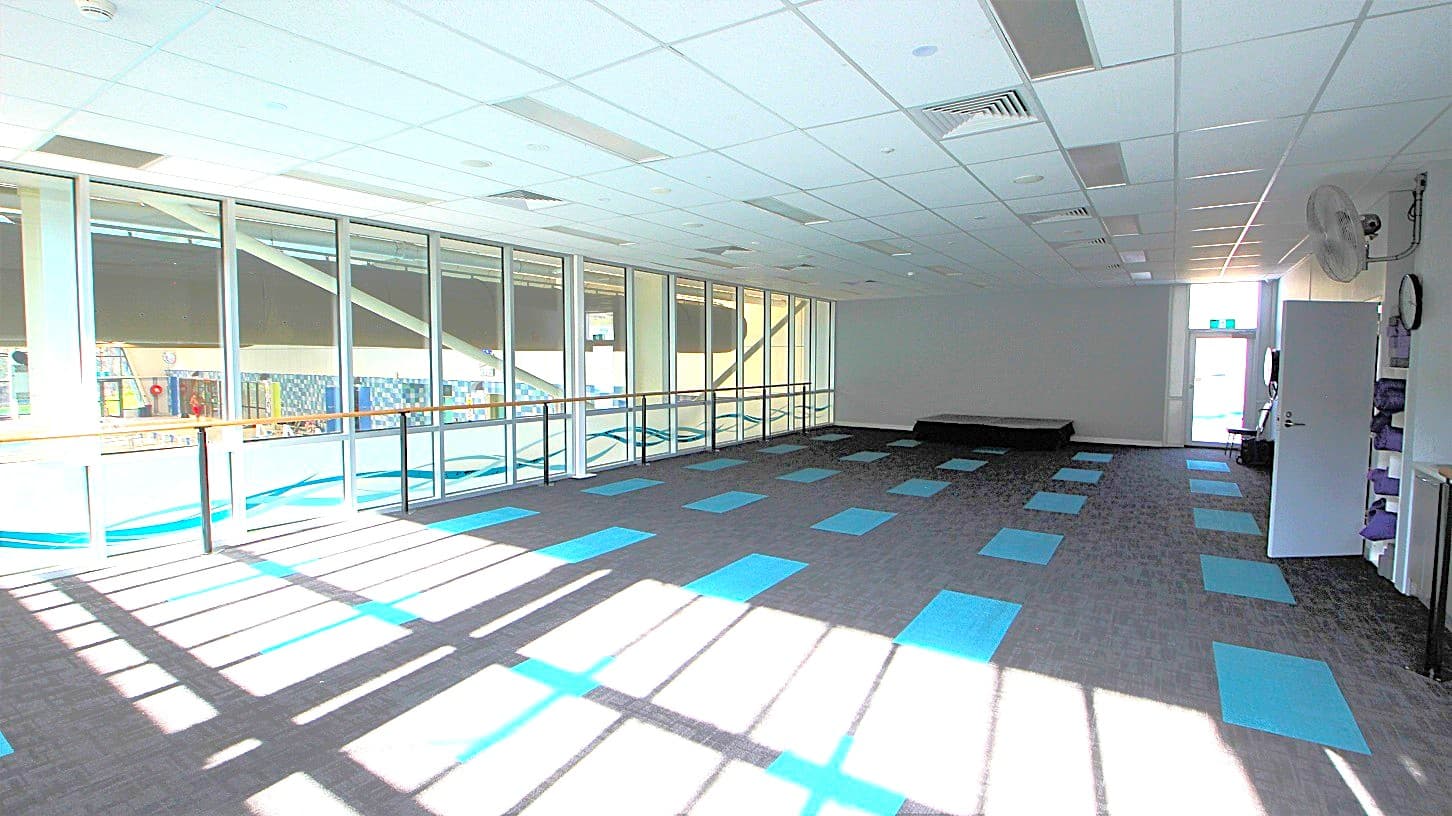 A well-ventilated yoga studio with glass walls, grey and blue rectangular carpet tiles, and wooden platforms for handheld during yoga practice. The front wall is painted grey while the ceilings are purely white.