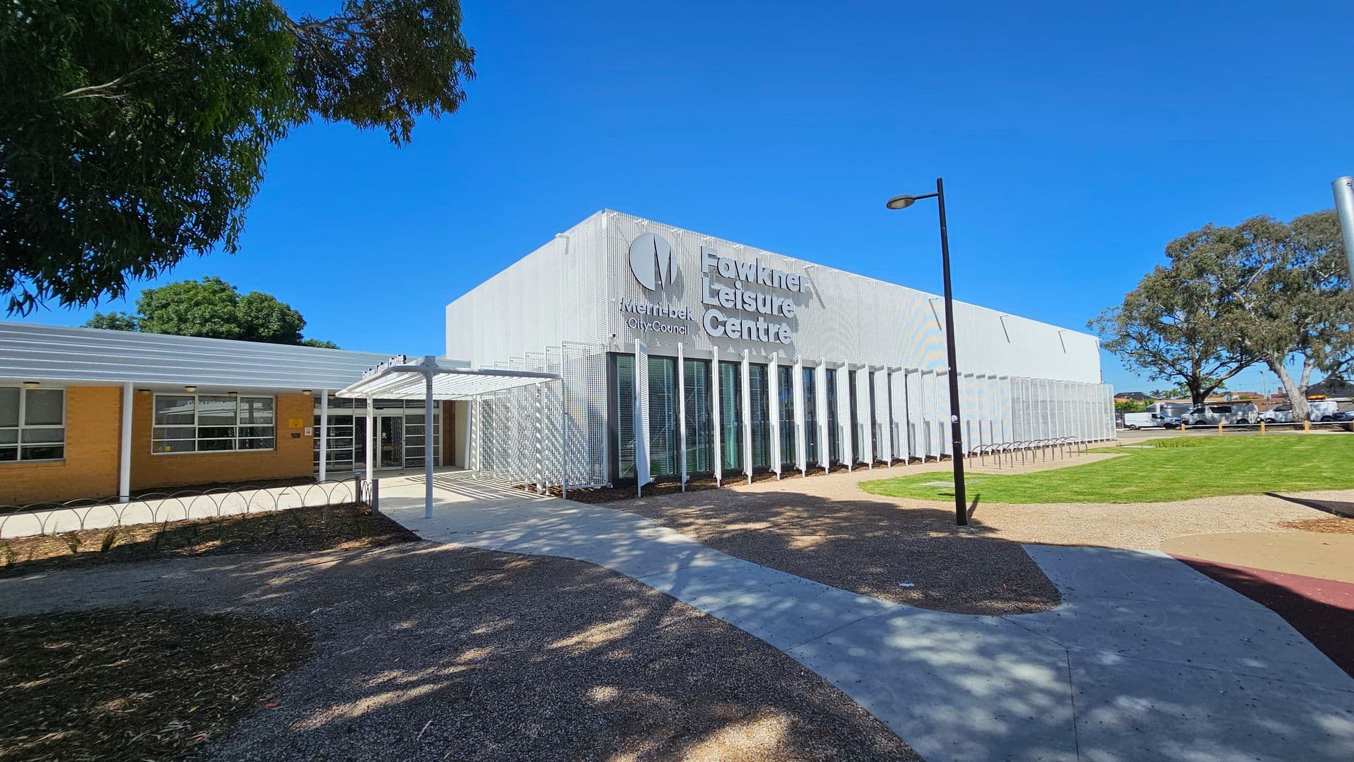 A modern building with a white facade and vertical slats on the right, labelled "Fawkner Leisure Centre" and "Merri-bek City Council". A pathway leads to the entrance, covered by a white metal canopy. On the left, a single-storey brick structure with large windows. A tree provides shade on the left, while a grassy area and a few parked cars are visible on the right. A tall streetlamp stands near the path.