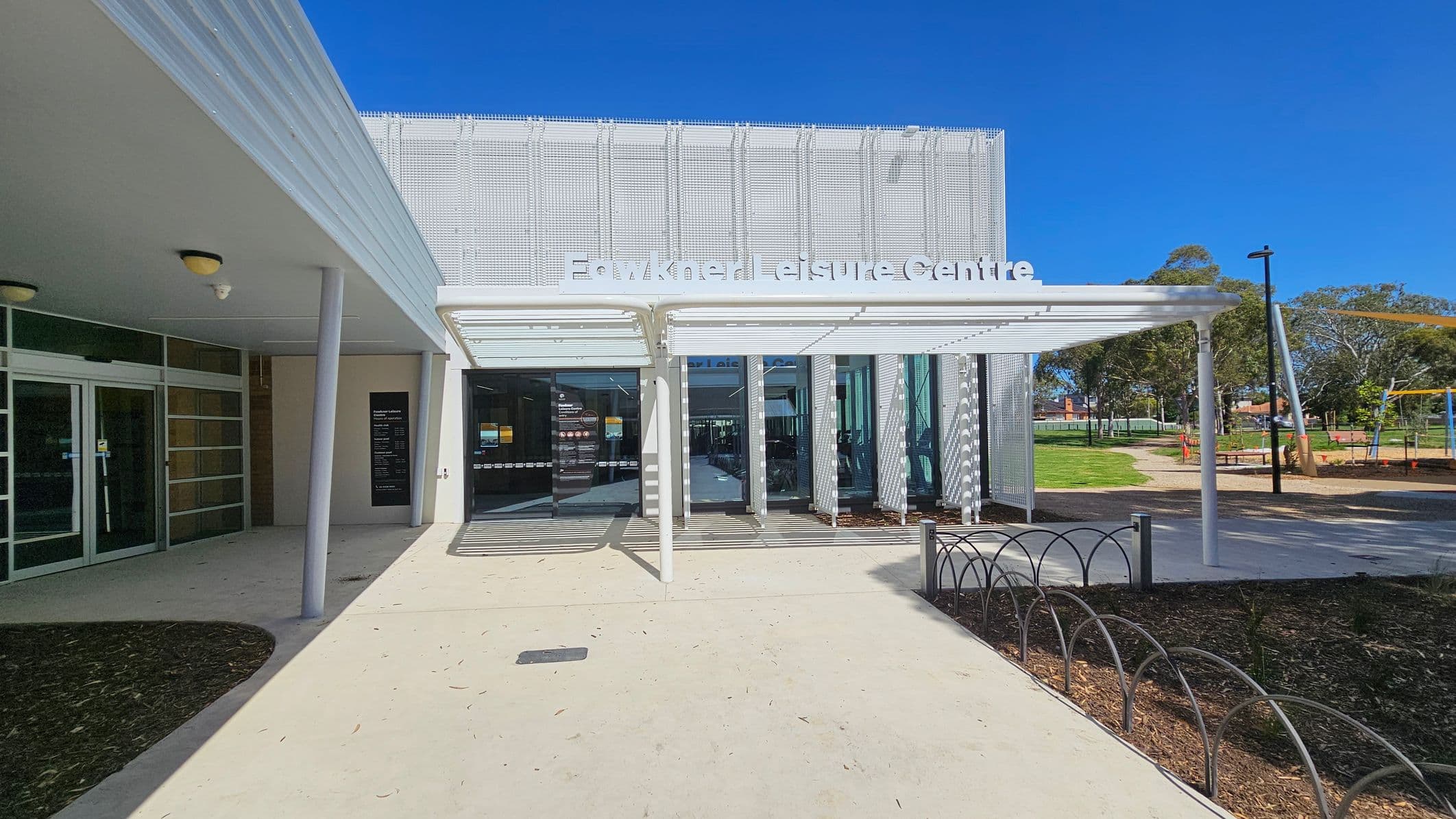 Entrance to a modern building with a sign reading "Fawkner Leisure Centre" above the door. The left side features glass doors and a covered walkway with a white roof supported by pillars. On the right, there's a grassy area with trees and a playground. The ground is paved, and there's a metal bike rack near the entrance.