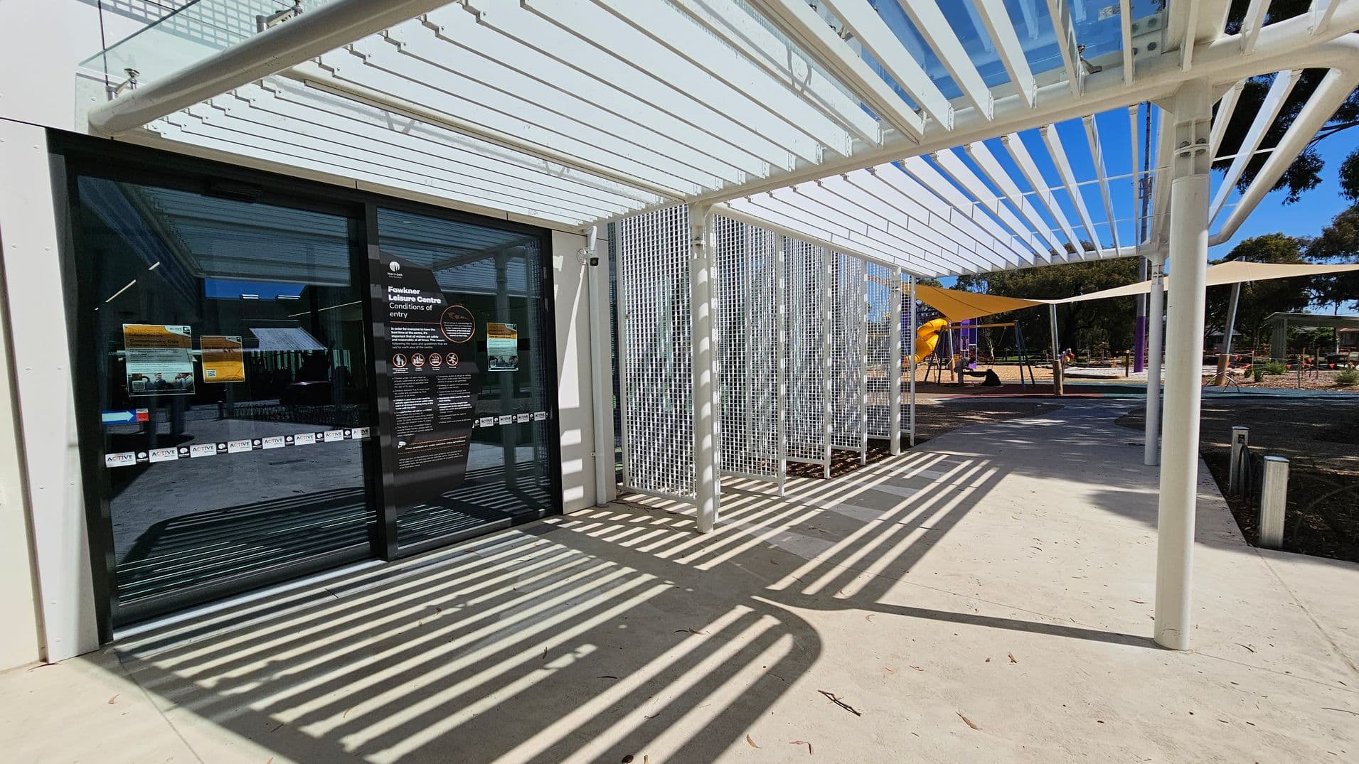 A modern entrance with glass doors on the left, displaying signage for the Fawkner Leisure Centre. The floor is light-coloured concrete with shadows from a white pergola above. To the right, a playground with yellow and purple equipment is visible, partially shaded by triangular fabric canopies. Trees and a bicycle are in the background.
