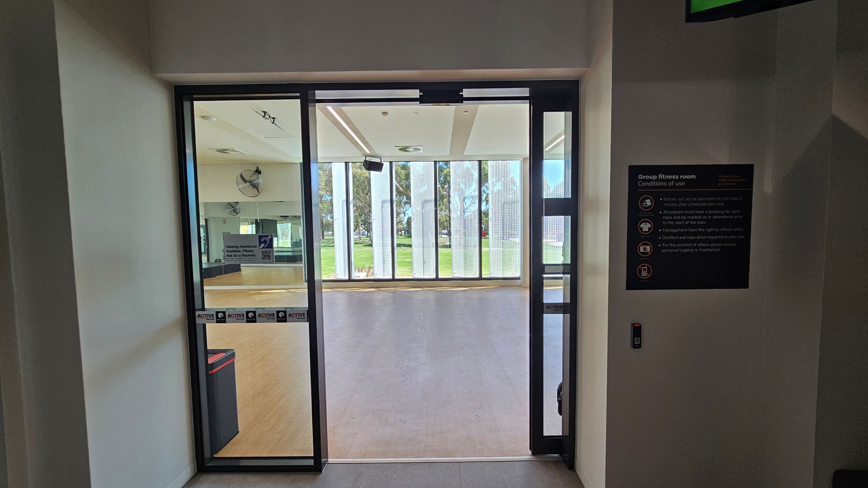 Glass doors leading to a group fitness room with a wooden floor. On the left, a sign with hearing assistance information is visible. The right side has a black sign listing the conditions of use for the room. Inside, a wall-mounted fan and large windows with vertical blinds overlook a grassy area outside. The ceiling has recessed lighting and a speaker. A small bin is visible near the door.