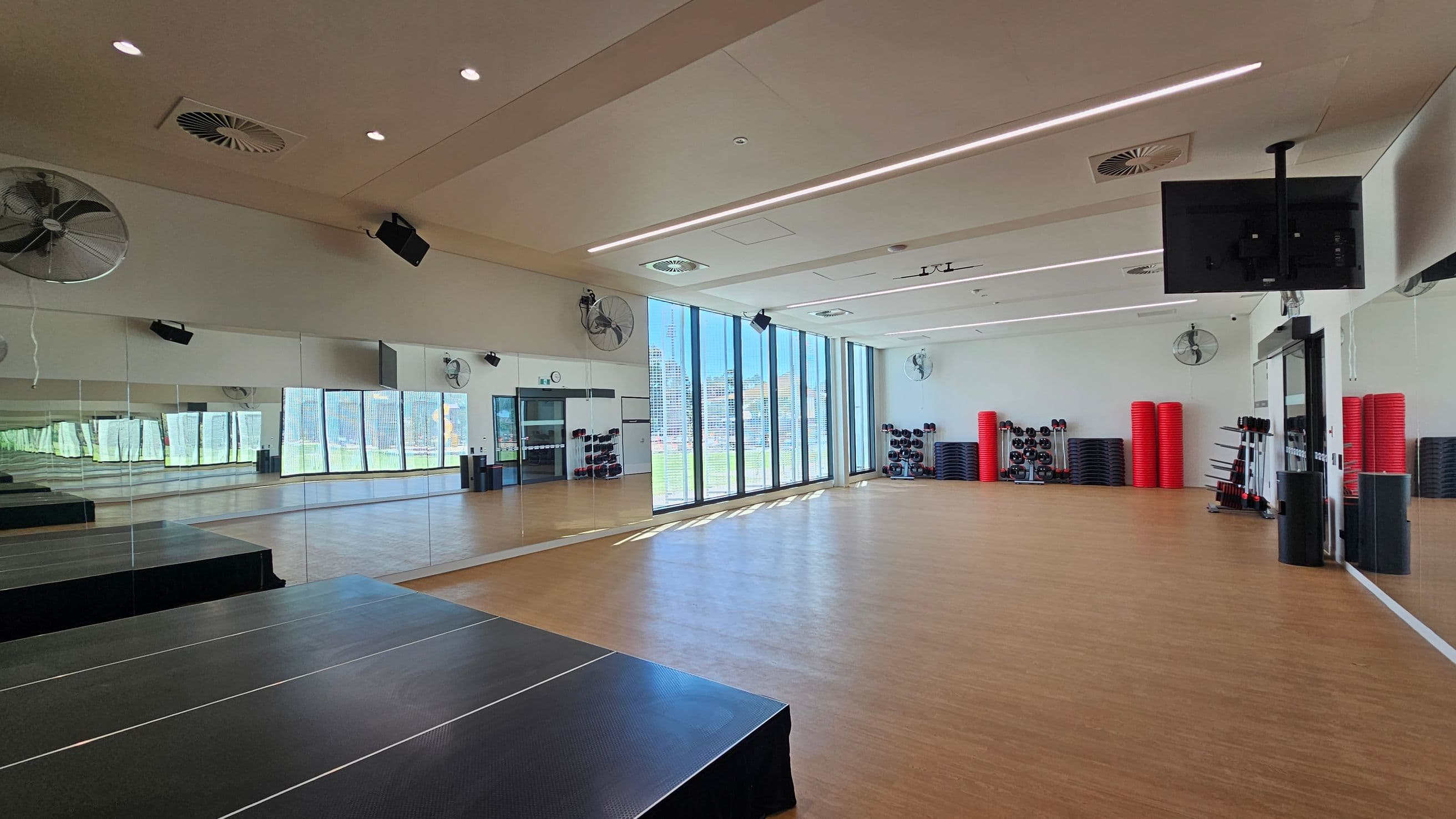 A spacious gym studio with wooden flooring and a mirrored wall on the left. Large windows on the right let in natural light. The back wall has stacks of red exercise mats and black weights. Ceiling fans and lights are visible above, and a black TV screen is mounted near the ceiling. A small stage is in the foreground on the left.