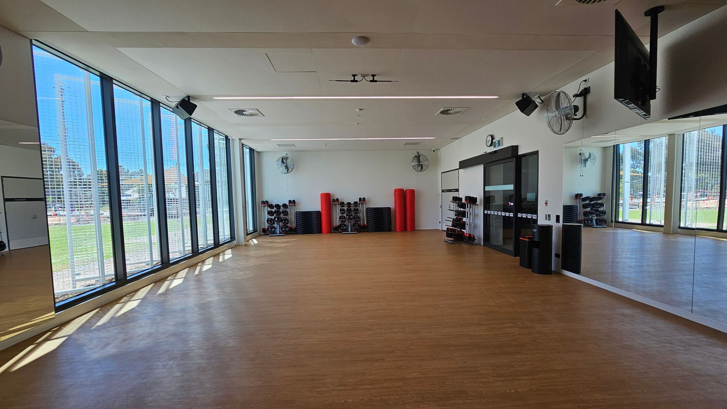 A spacious fitness studio with a wooden floor and a white ceiling. On the left, large windows allow natural light to flood in, offering a view of the outdoors. The back wall has stacks of red exercise mats and black weights. On the right, a mirrored wall reflects the room, and a mounted TV is visible. Ceiling fans and speakers are installed above.
