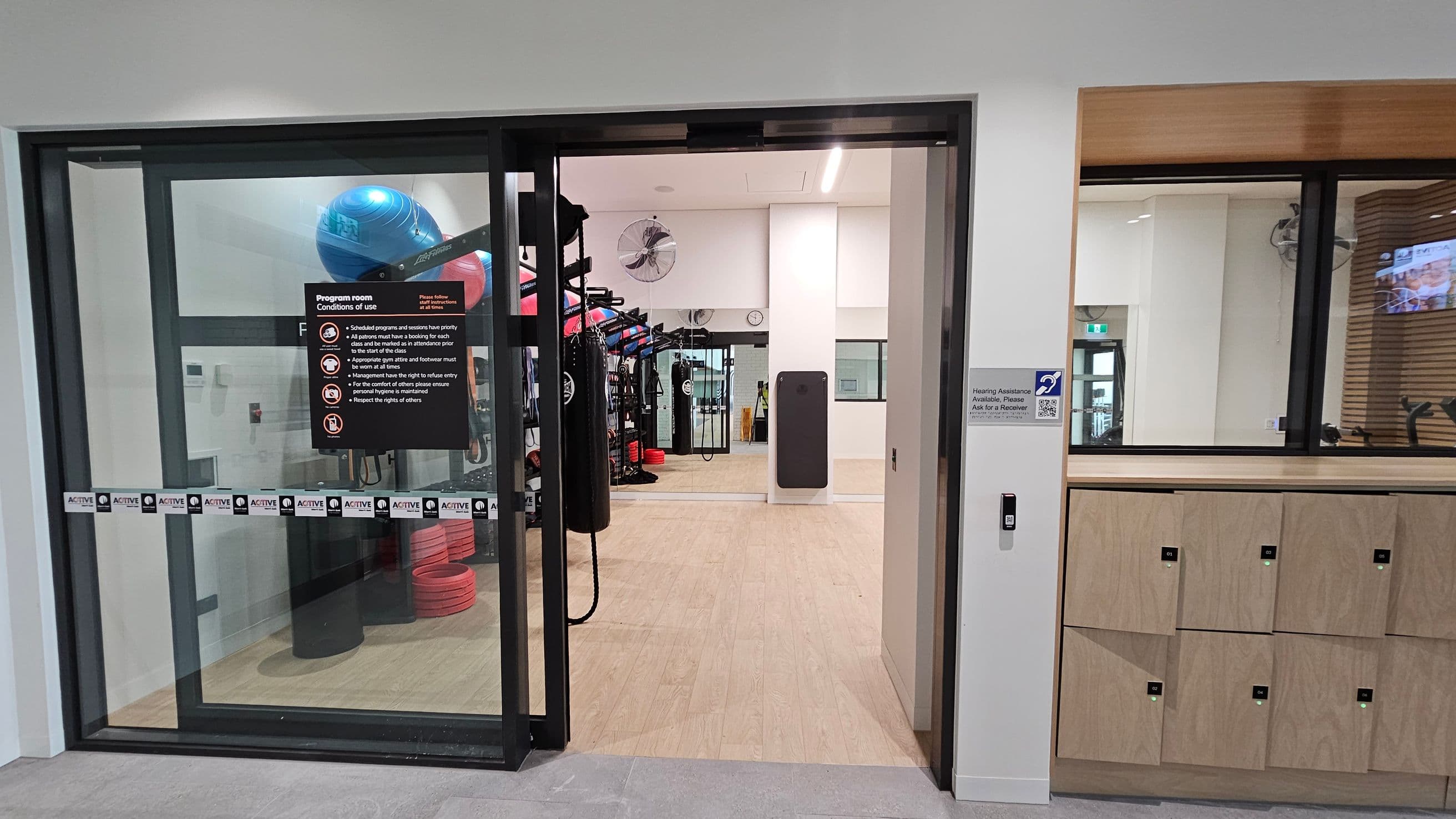 Glass doors lead into a gym room. On the left, a sign lists the conditions of use. Inside, exercise equipment like punching bags and stacked red steps are visible. The floor is light wood, and the ceiling has recessed lighting. On the right, there are wooden lockers and a sign indicating hearing assistance availability. A wall-mounted fan and a clock are visible in the background.
